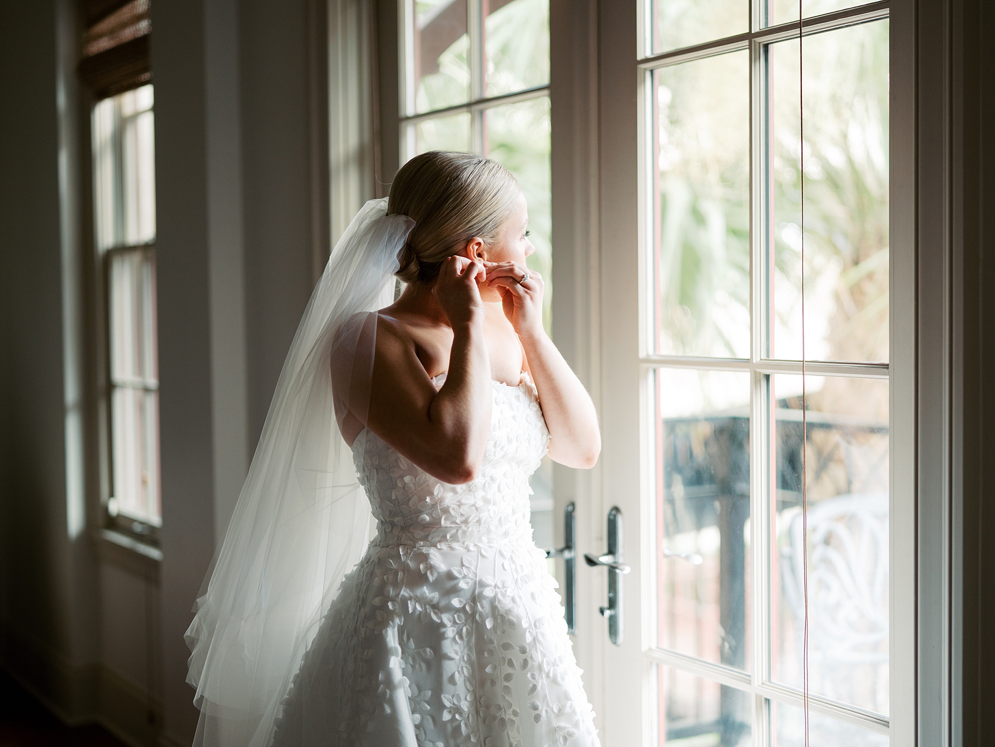 bride puts on earrings in window of home wearing Anne Barge wedding dress with textured bodice and skirt