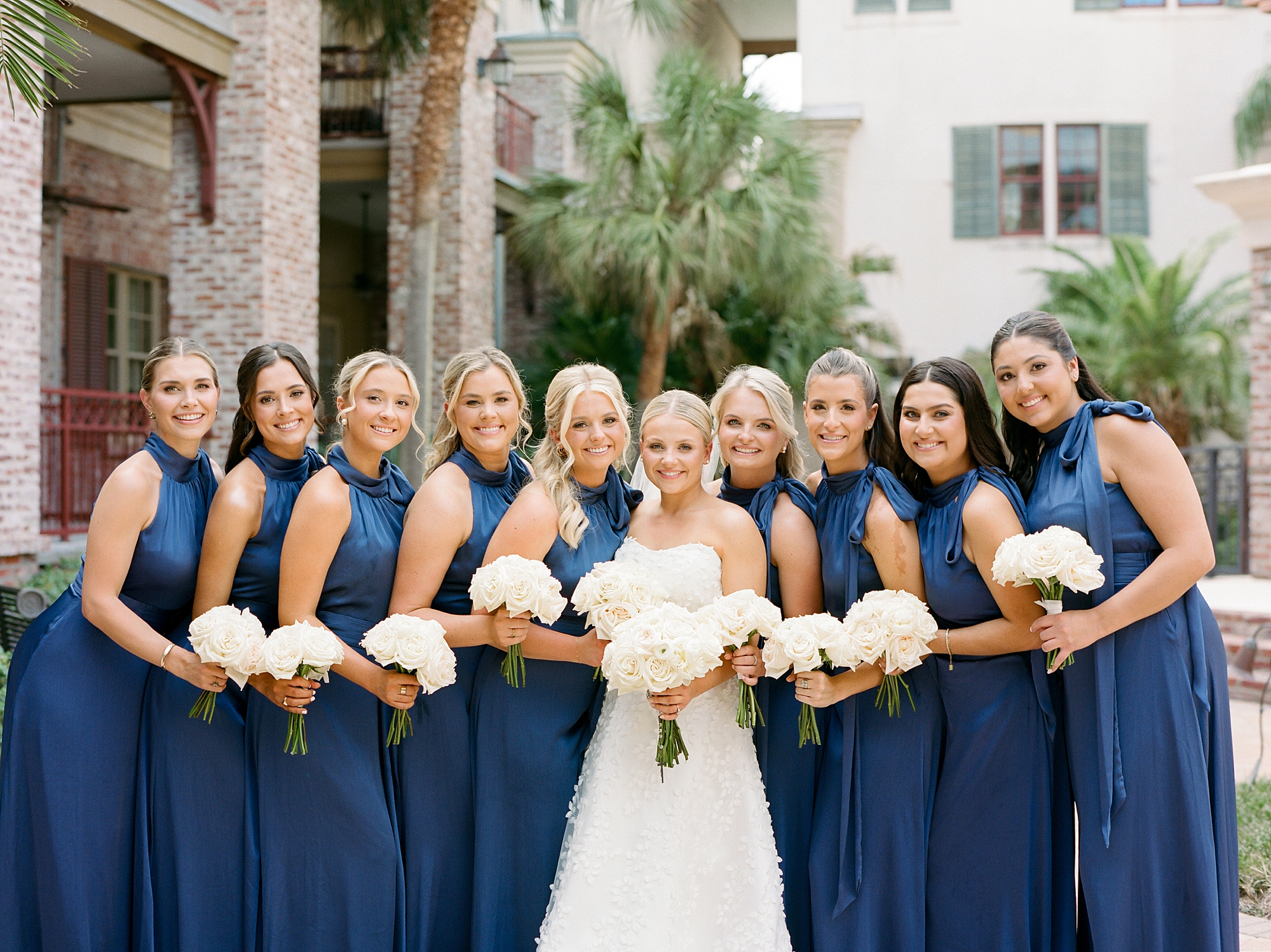 9 bridesmaids in one-shouldered blue dresses hold bouquets of white flowers next to bride 
