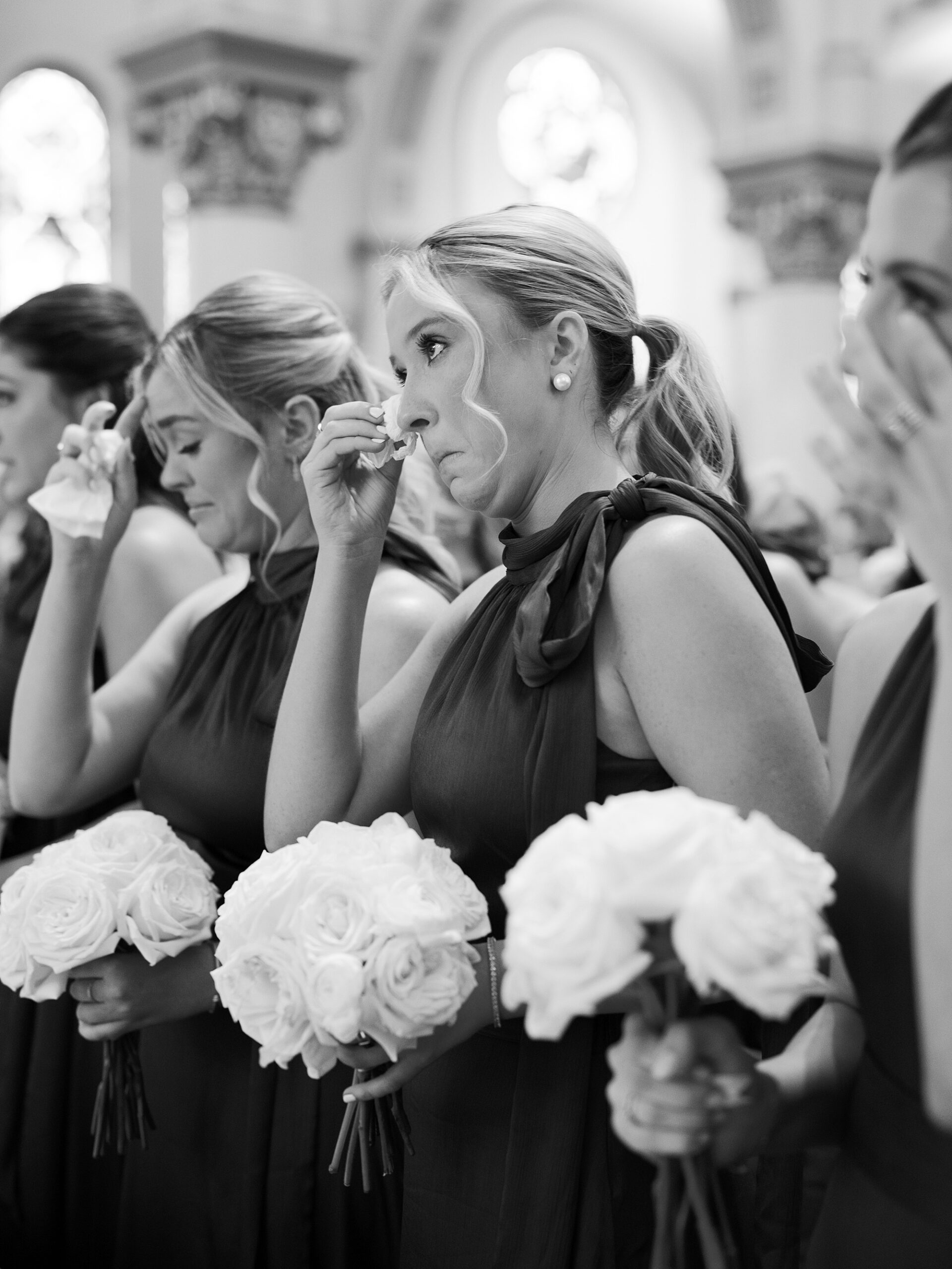 bridesmaids holding white bouquets wipe away tears from their eyes during traditional Catholic Church wedding at St. John Cathedral
