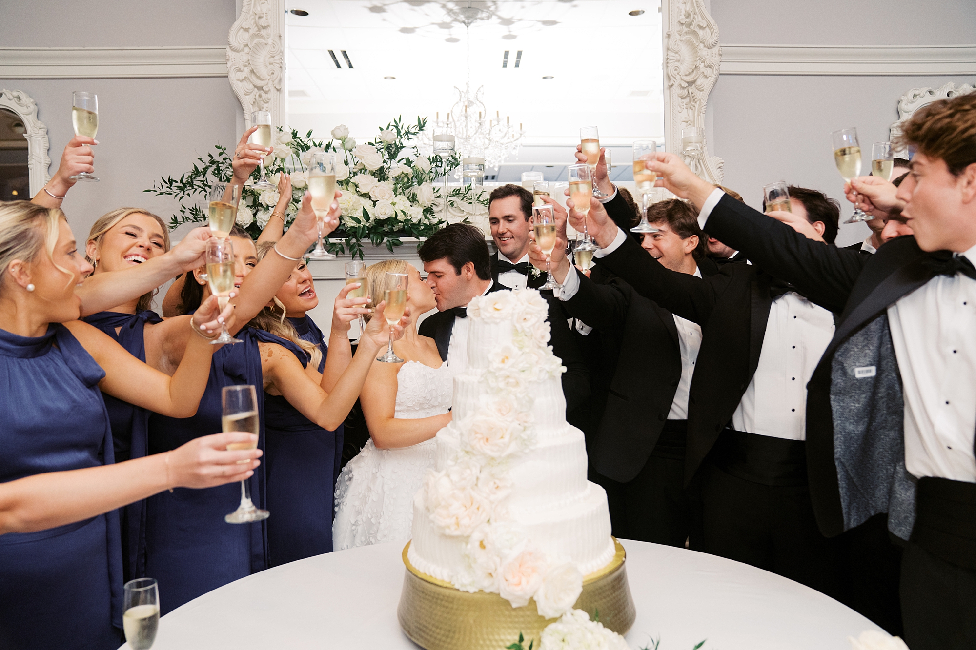 newlyweds kiss by tiered wedding cake with wedding party cheering around them