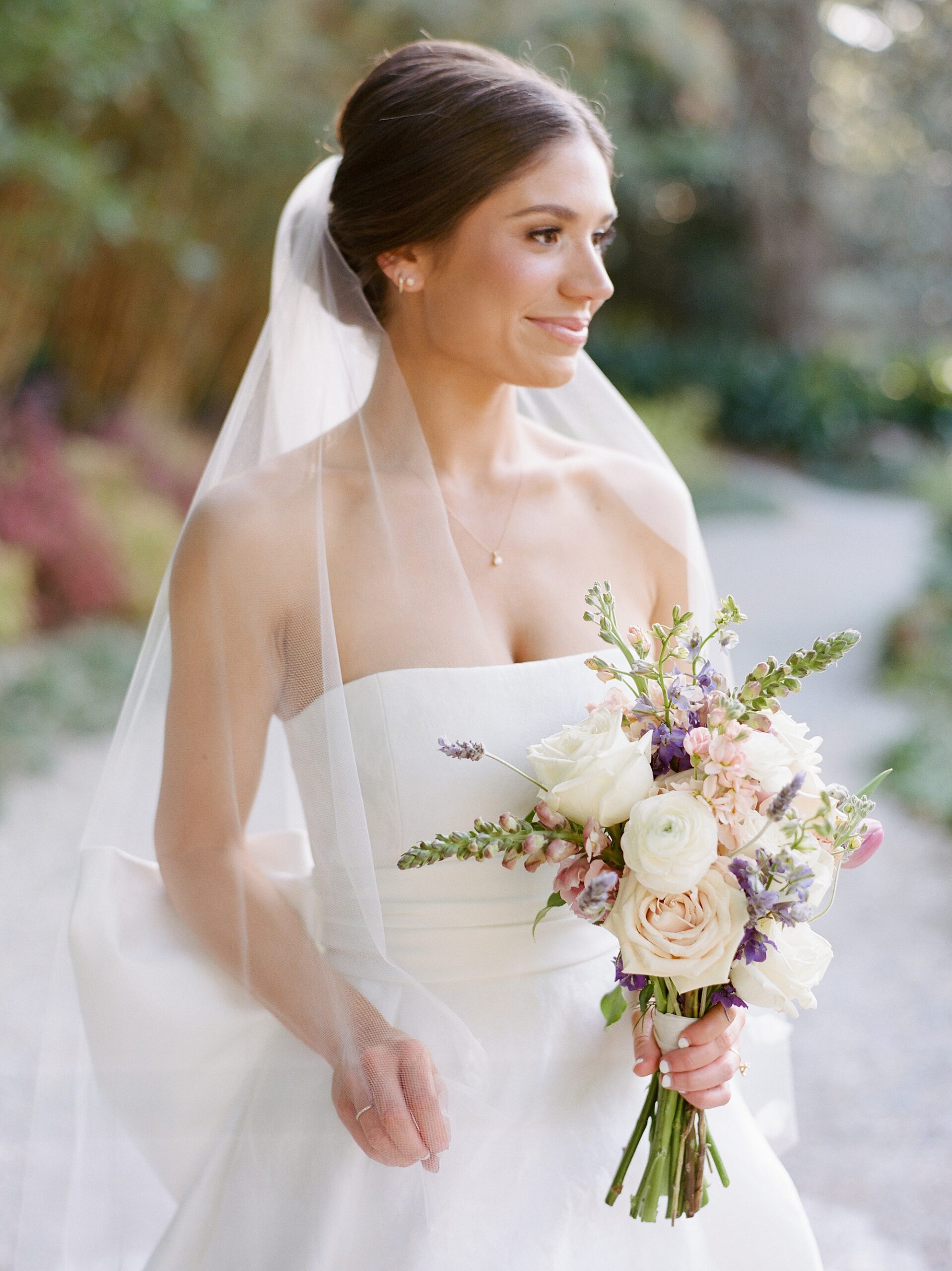 bride smiles with veil around her shoulders in strapless wedding gown with white, pink and purple flowers 
