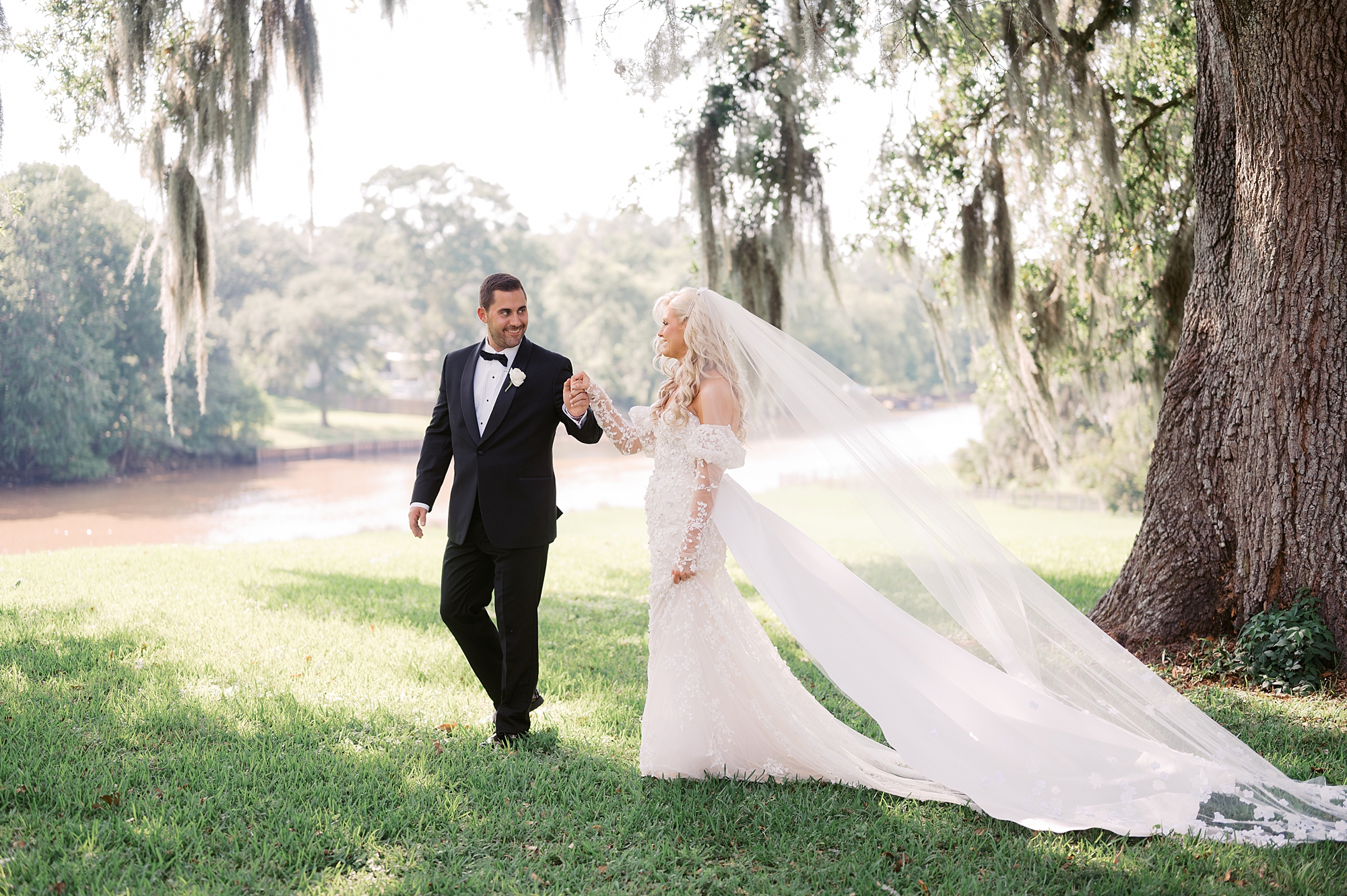 groom leads bride across lawn under tree with Spanish moss 