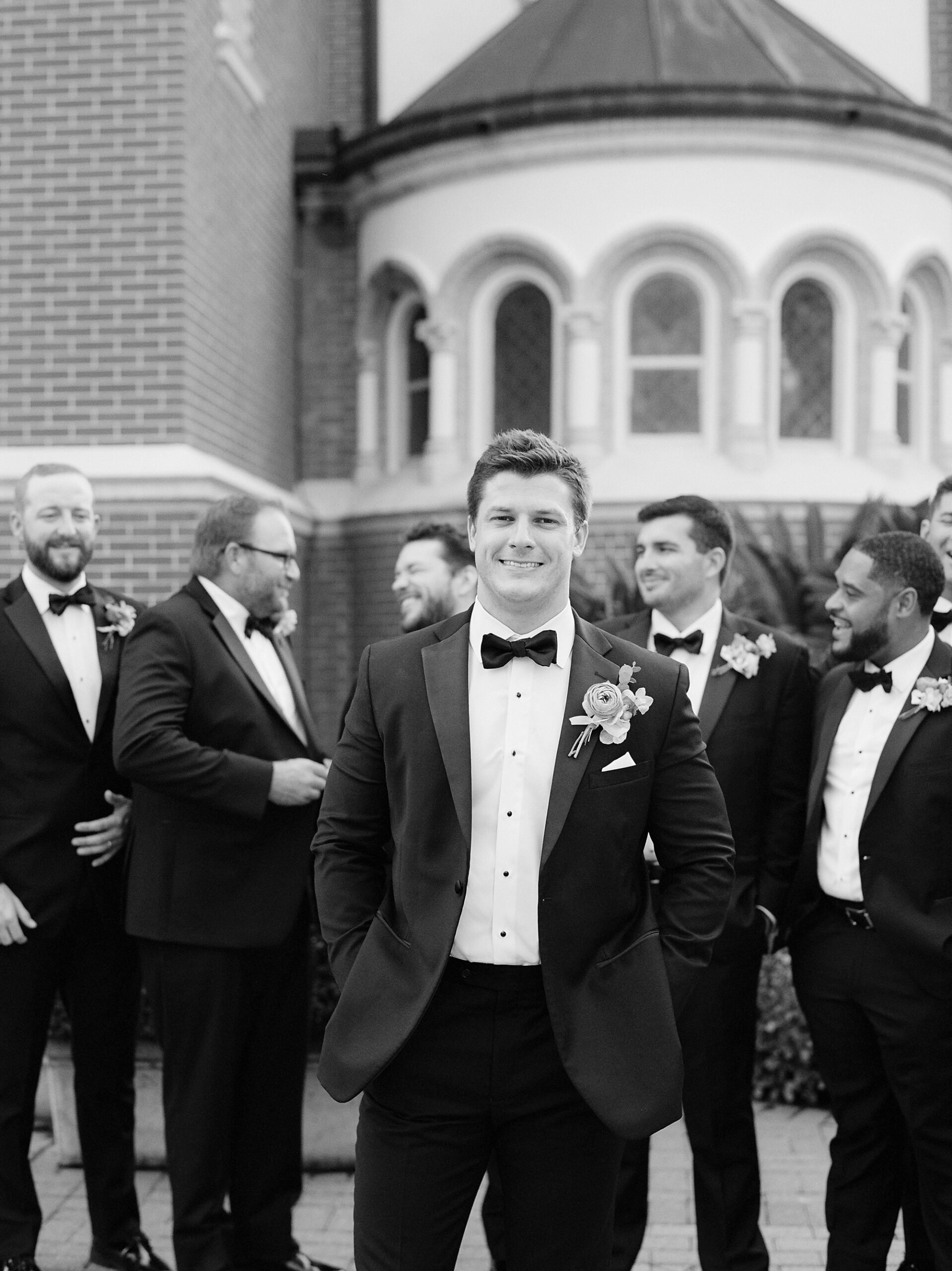 groom in black tux walks in front of groomsmen outside the Cathedral of St. John the Evangelist
