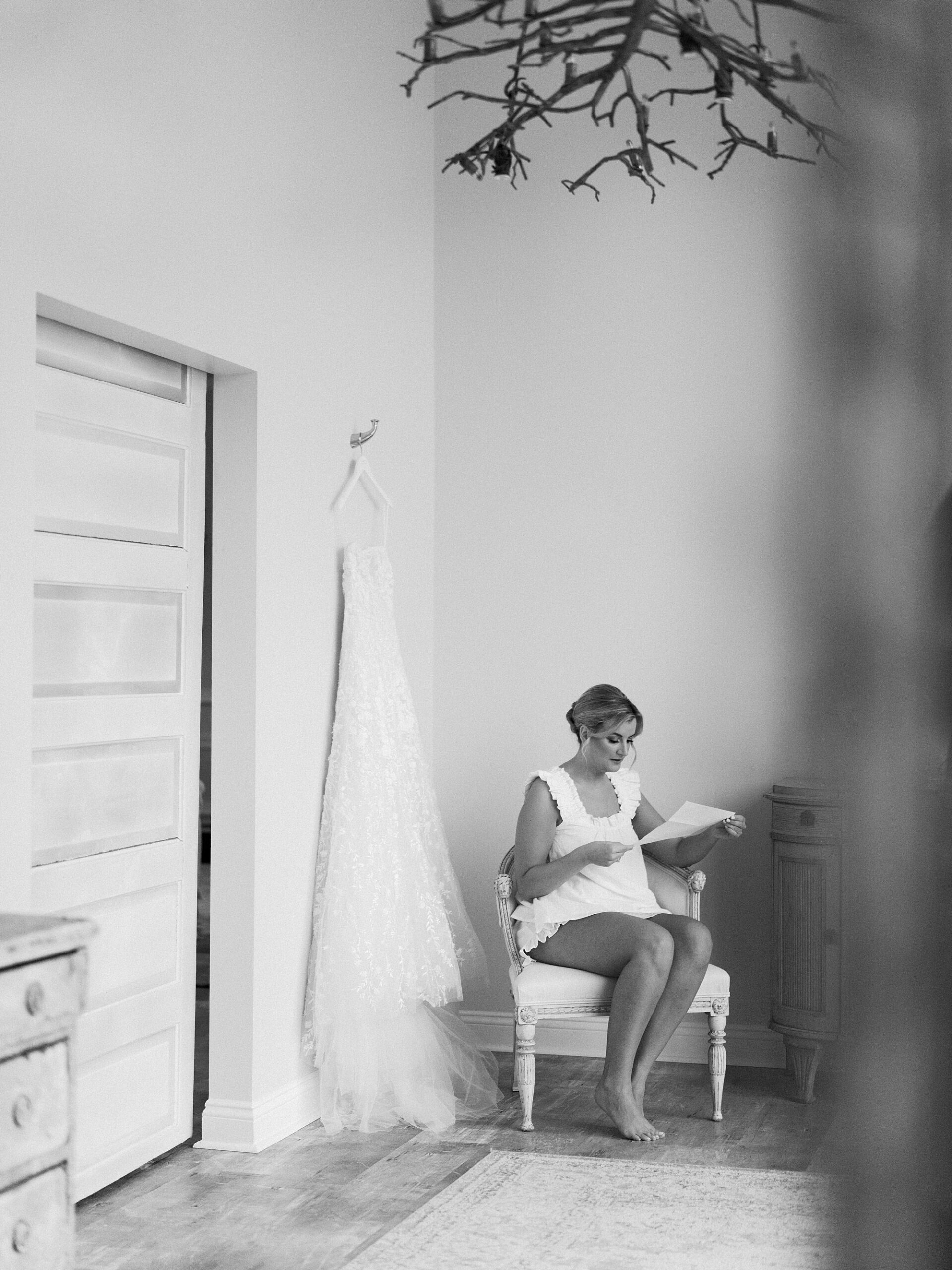 bride sits reading a letter by wedding dress hanging on wall 