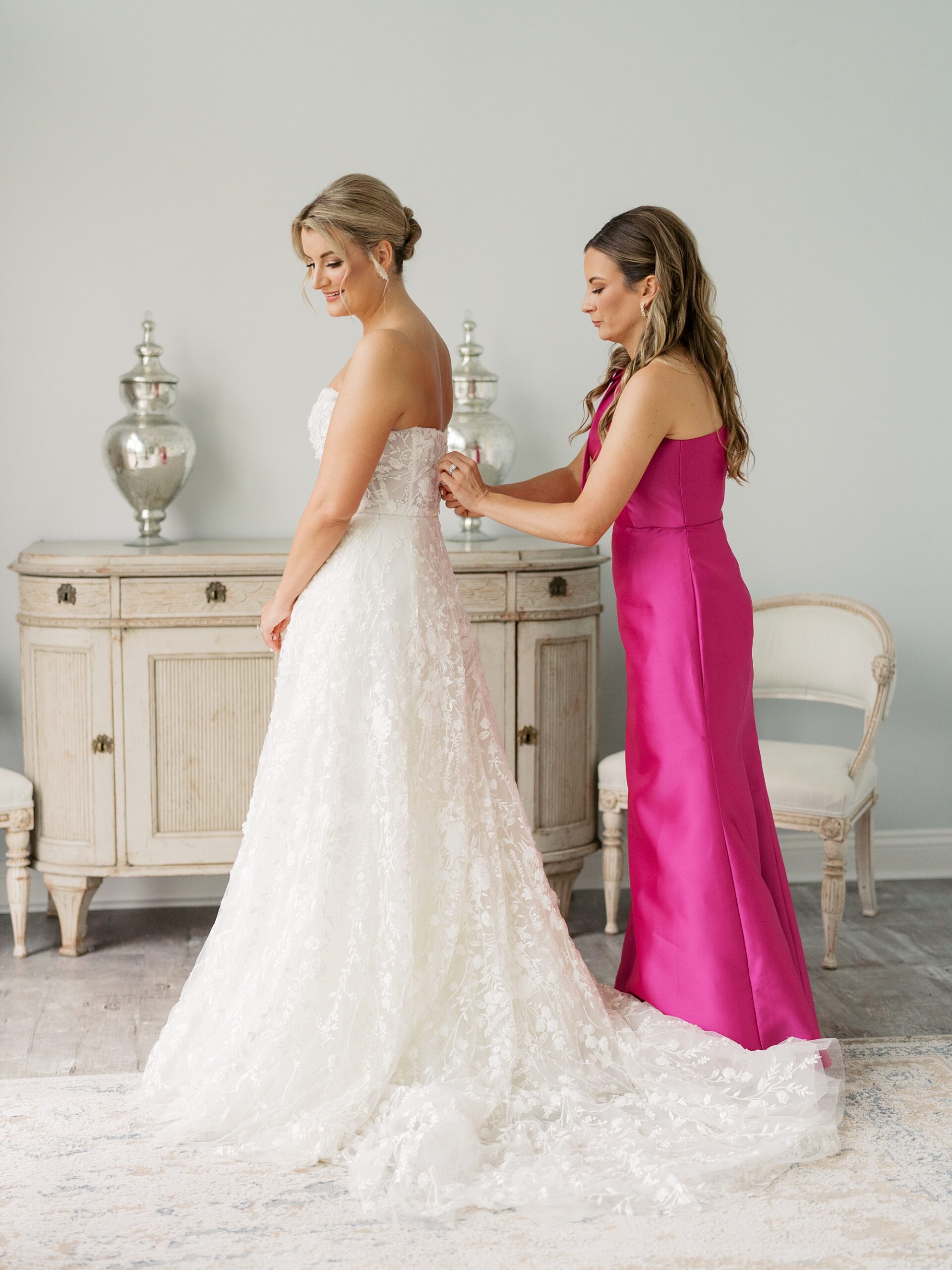 bridesmaid in bright pink gown helps bride into wedding dress