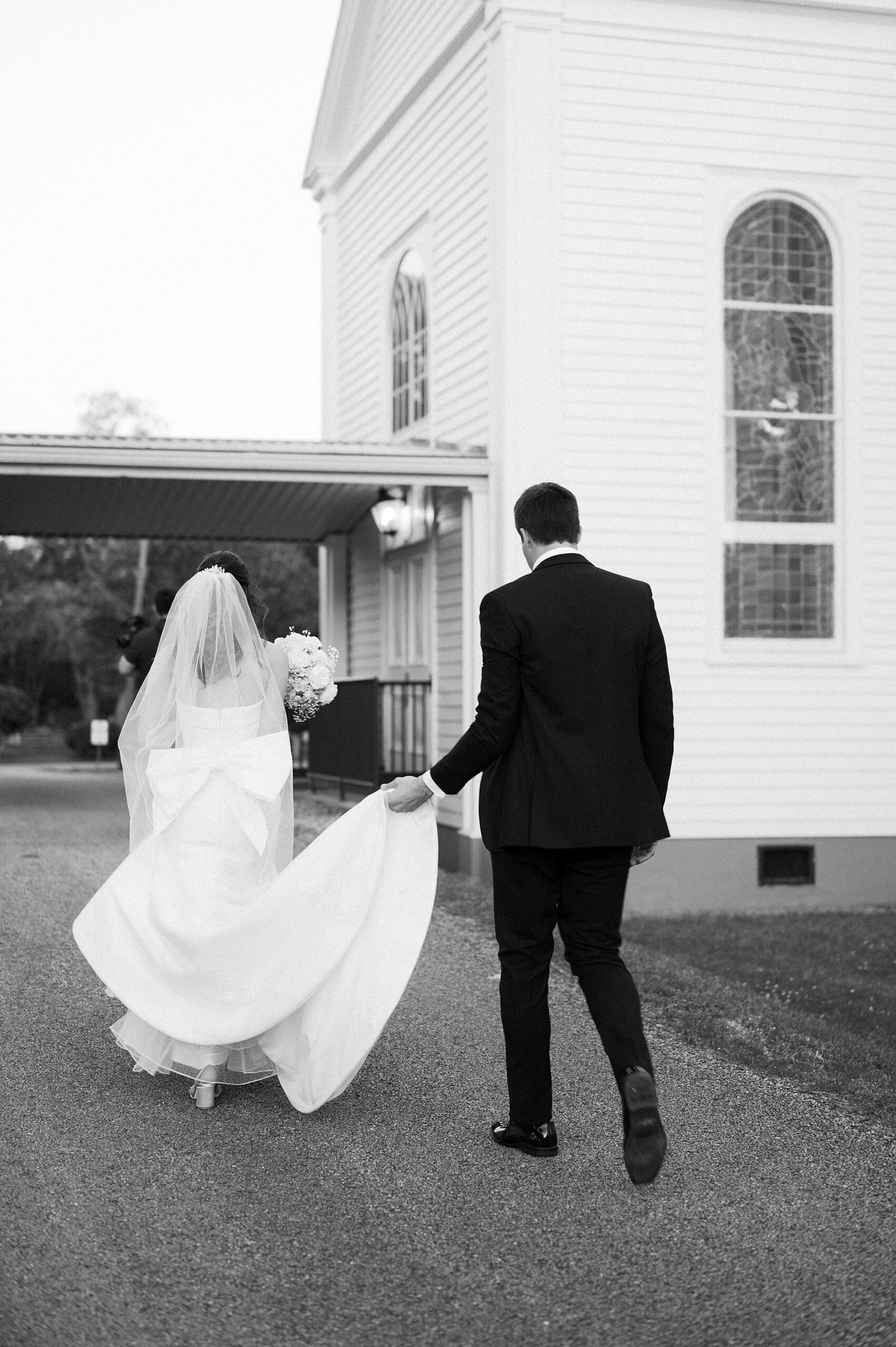 groom holds up back of bride's dress walking towards St. Charles Borromeo