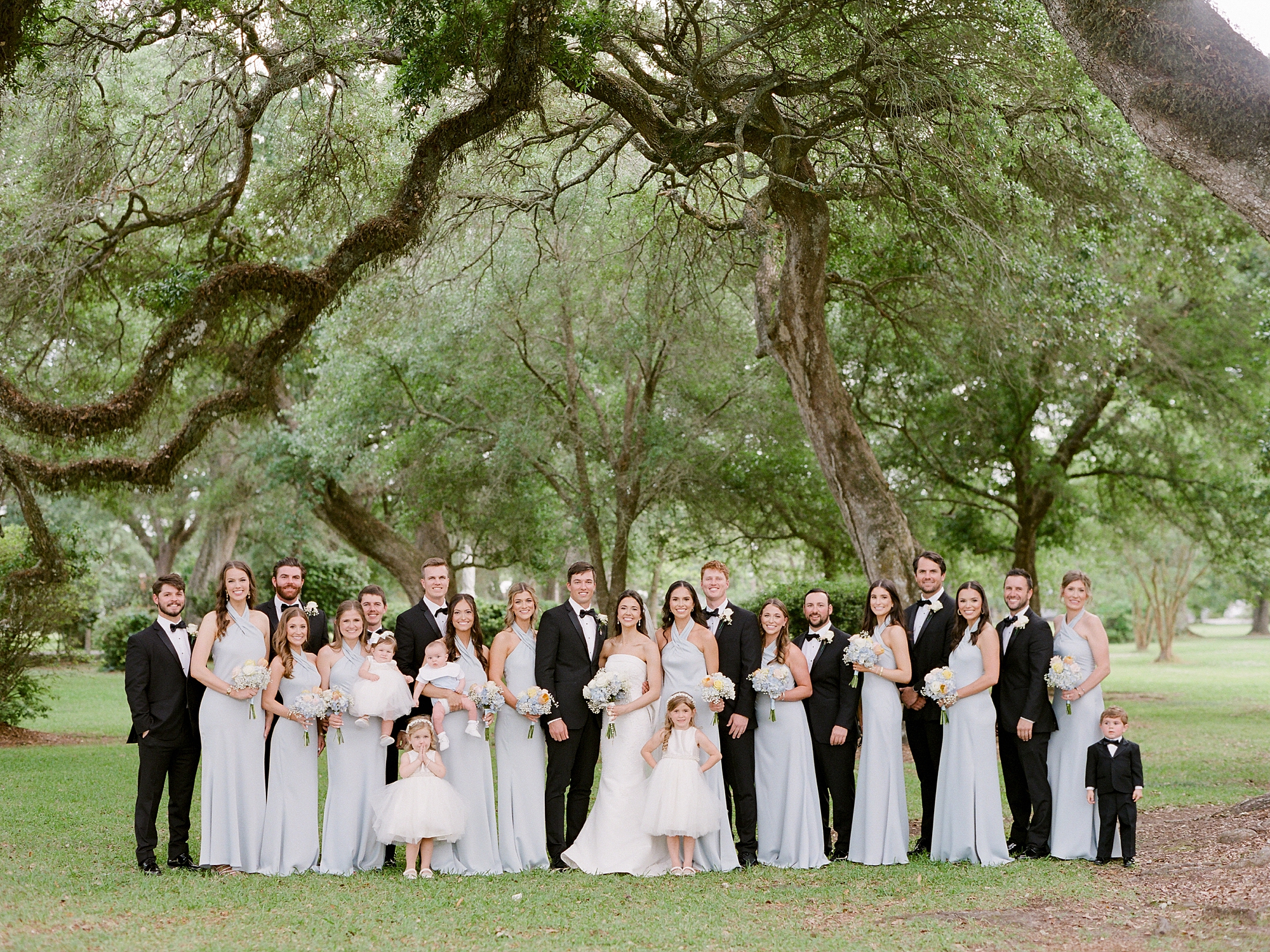 bride and groom stand with wedding party under oak trees outside St. Charles Borromeo