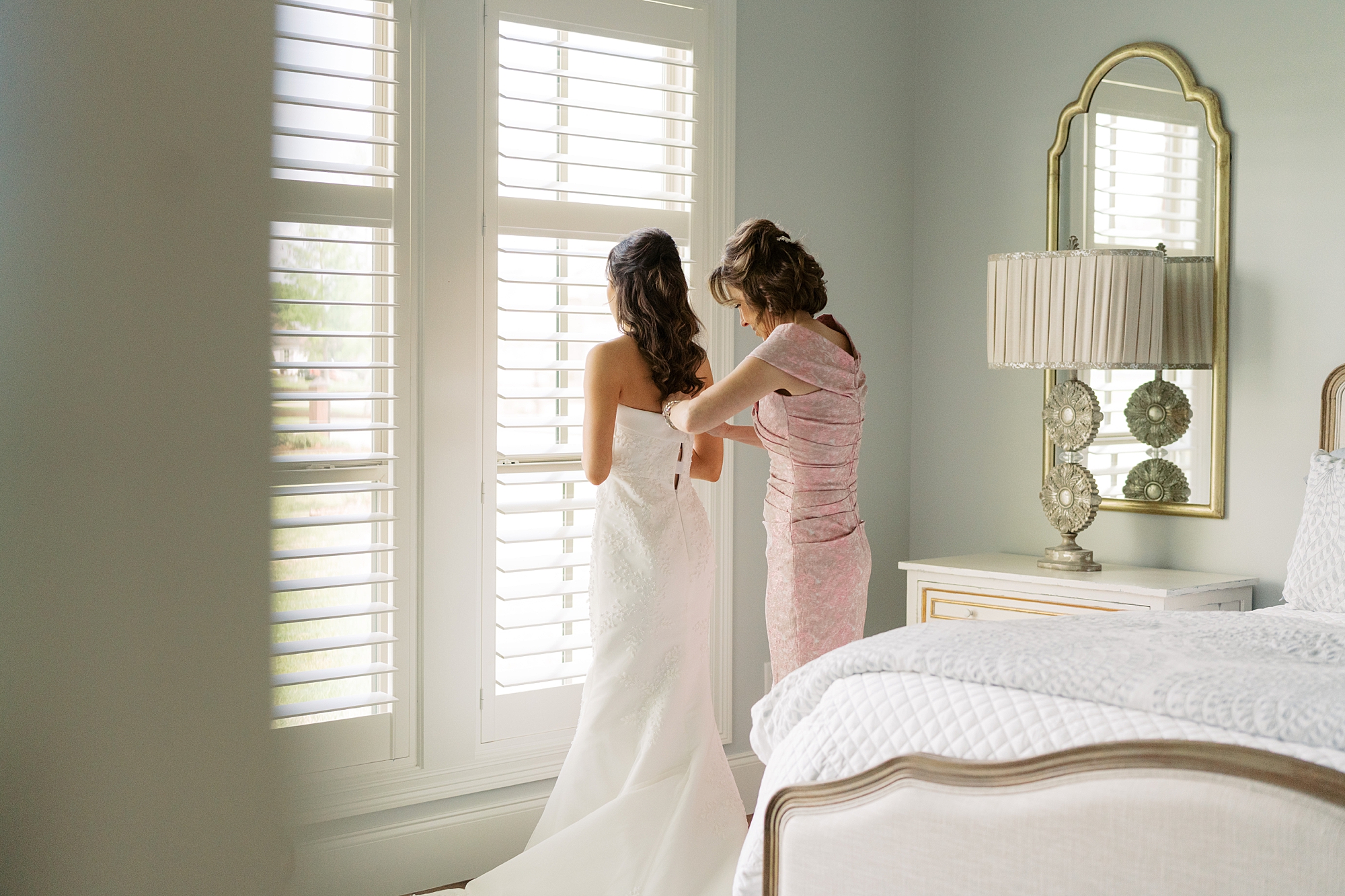 mother in pink gown helps bride with wedding dress on morning of Lafayette LA wedding day