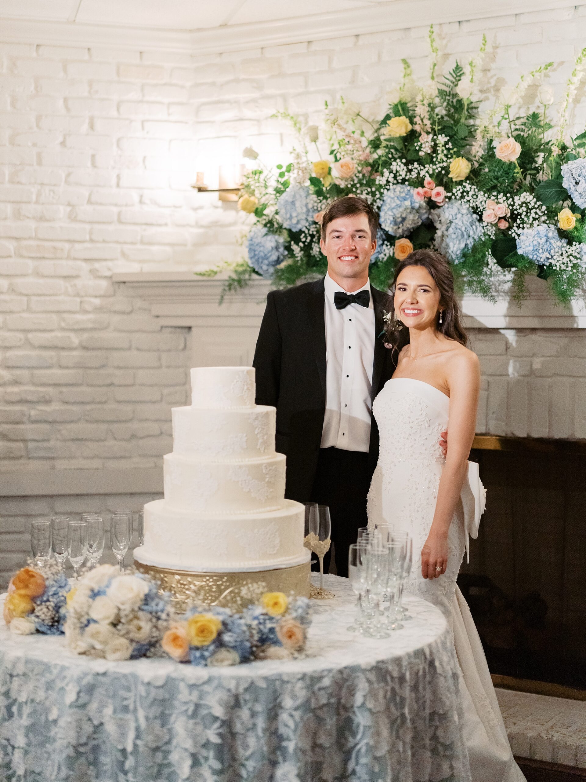 newlyweds stand by cake in front of fireplace with blue floral arrangement 