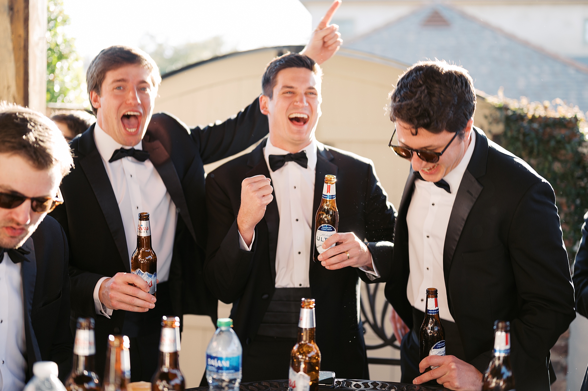 groom laughs with three groomsmen drinking beers on morning of wedding 