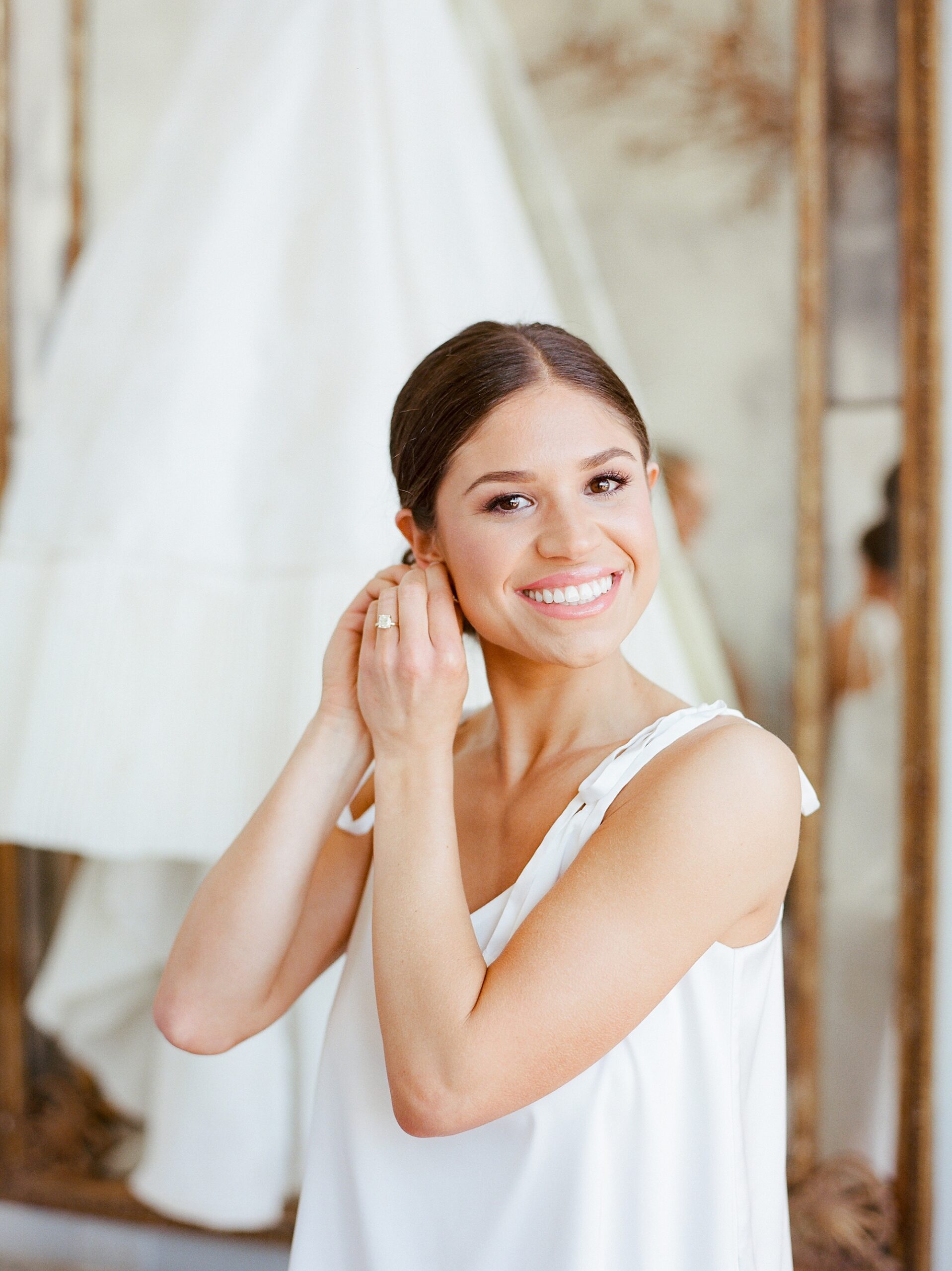 bride adjusts earrings on morning of wedding in front of gown 