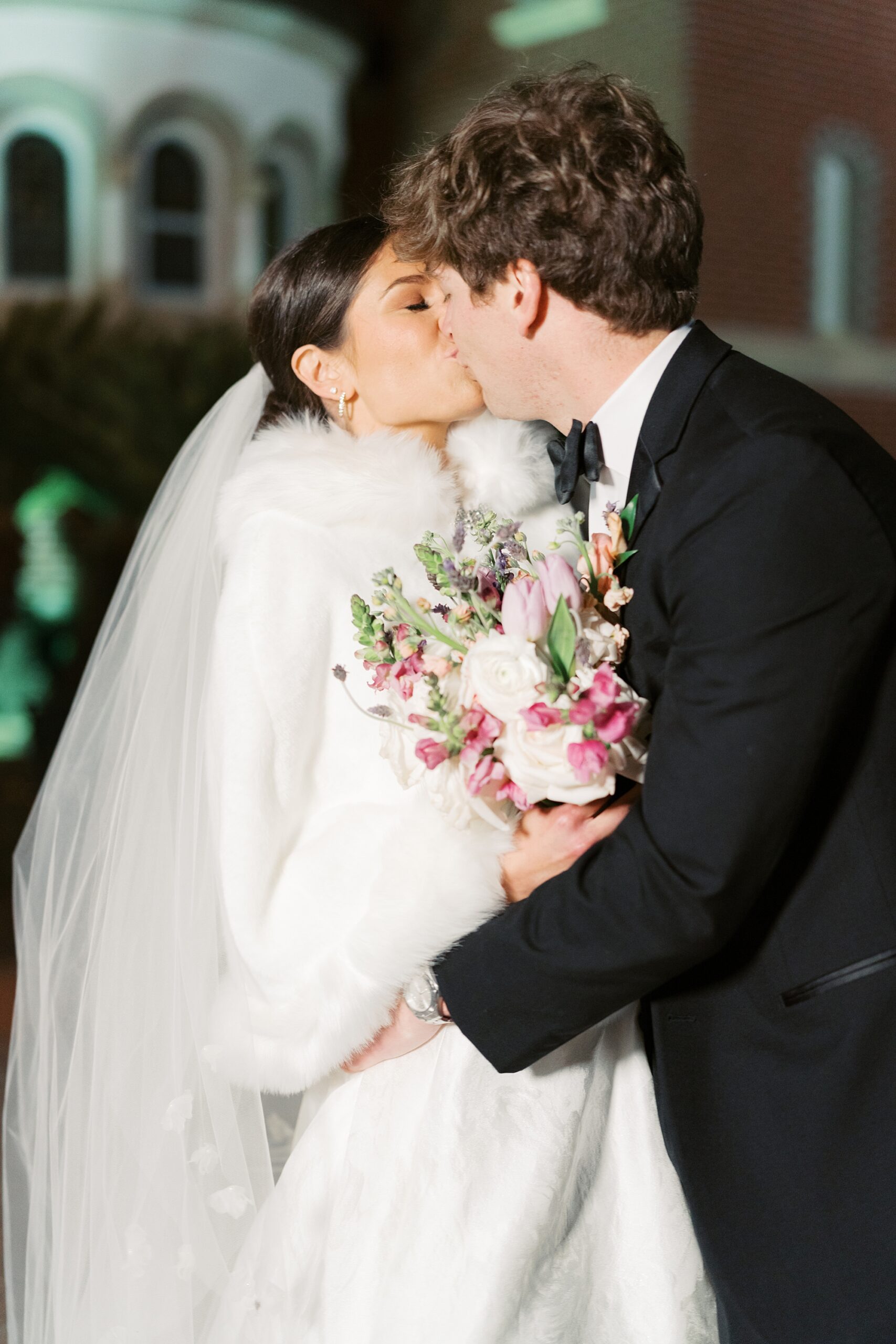 groom kisses bride in dress with fur around her shoulders 
